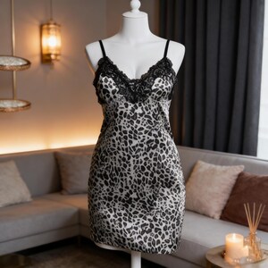 Op de afbeelding: Een slipdress met luipaardprint op een mannequin. De jurk heeft zwarte kanten afwerking langs de halslijn en dunne zwarte bandjes. De stof heeft een patroon van zwarte vlekken op een crèmekleurige achtergrond. De jurk is kort en eindigt boven de knieën.