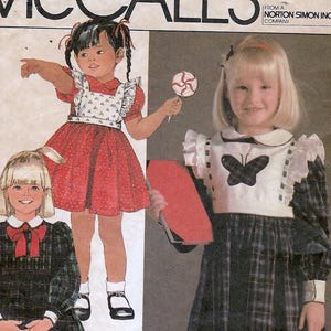 May include: McCall's sewing pattern for children's dresses. The pattern features illustrations of girls wearing dresses with ruffles, lace, and plaid patterns. The text on the pattern reads "Ruffles & Lace" and "Child Size 2".