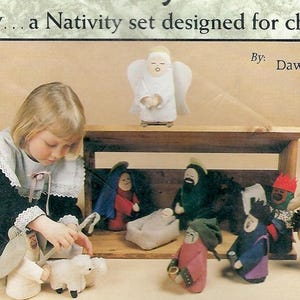 May include: A nativity set featuring fabric figures including an angel, a manger, and various characters. The set includes a camel, sheep, and human figures in colourful clothing. The text reads: Finally... a Nativity set designed for children! By: Dawn Fuchs.