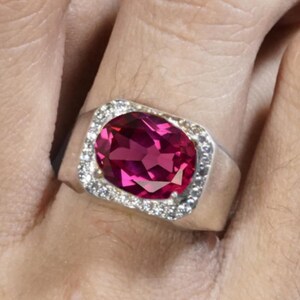 Op de afbeelding: Een zilveren ring met een grote, ovaal geslepen, dieproze edelsteen. De edelsteen is omgeven door een halo van kleinere, heldere stenen. De ring is gezet op een brede band.