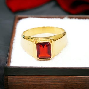 Könnte beinhalten: Ein goldener Ring mit einem rechteckigen, roten Edelstein. Der Ring liegt auf einer weißen Oberfläche in einer Holzbox. Das Ringband ist dick und poliert. Der Edelstein hat eine leuchtend rote Farbe.