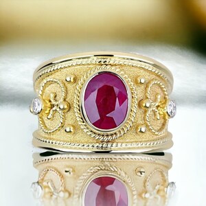 Può includere: Un anello d'oro con una grande pietra centrale ovale di rubino. L'anello ha una fascia strutturata con elementi decorativi e due pietre più piccole. Il rubino è di un colore rosso intenso e l'anello è molto riflettente.