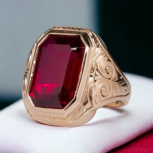 May include: A gold ring with a large, rectangular, deep red gemstone. The ring has an ornate, engraved design around the gemstone and on the band. The ring is set on a white surface.