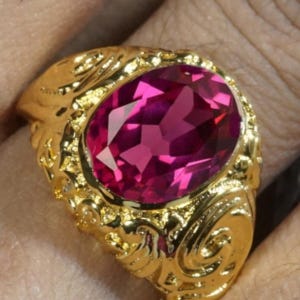 Könnte beinhalten: Ein goldener Ring mit einem ovalen, facettierten, magentafarbenen Edelstein. Der Ring hat ein verziertes, strukturiertes Goldband mit einem Wirbeldesign um den Edelstein. Der Ring ist ein auffälliges Schmuckstück.