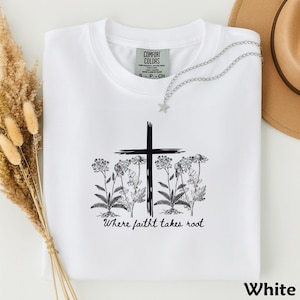 May include: A white t-shirt featuring a black graphic of a cross surrounded by wildflowers. Below the graphic, the text reads "Where faitht takes root". The shirt has a Comfort Colors label at the neck and a silver-toned star charm necklace.