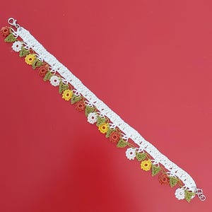 May include: A handcrafted crochet bracelet featuring a white base band adorned with a repeating pattern of small, colorful flowers in white, yellow, and rust orange, interspersed with tiny green leaves and finished with a silver-toned metal lobster clasp.