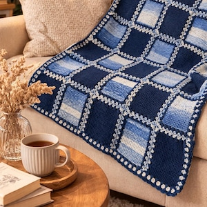 Puede incluir: Una manta de ganchillo azul y blanca hecha a mano con un patrón geométrico. Está colocada sobre un sofá beige. Una taza de té blanca y un libro están sobre una mesa auxiliar de madera. El diseño de la manta presenta cuadrados en tonos azules.