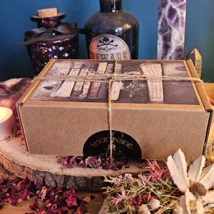 May include: A brown cardboard box tied with twine, displaying a vintage book design, with a sticker saying "MAGIC INSIDE". The scene includes potion bottles, a purple crystal, and dried flowers, all arranged on a wooden surface.
