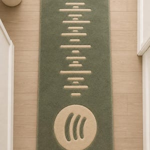 Pode incluir: Um tapete de banho retangular verde sálvia com um design de onda sonora bege e um logotipo circular. O tapete é feito de um material macio e texturizado e é colocado em um piso de madeira clara. O logotipo está no centro inferior.