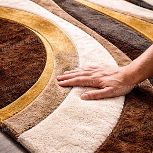 Könnte beinhalten: Nahaufnahme eines braunen, beigen und goldenen Teppichs mit einem wellenförmigen Muster. Der Teppich hat eine weiche, strukturierte Oberfläche. Die Worte "MODERNS WEAVE" sind auf dem Teppich aufgedruckt.