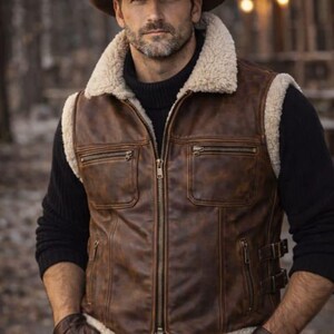 Könnte beinhalten: Eine braune Lederweste mit cremefarbenem Shearling-Futter an Kragen und Kanten. Die Weste hat einen Reißverschluss, zwei Brusttaschen und zwei untere Taschen. Die Person auf dem Bild trägt einen braunen Cowboyhut und Handschuhe.