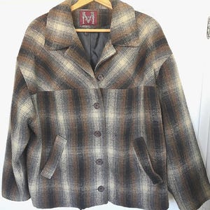 May include: A brown, tan, and black plaid jacket with a collared neckline, button closure, and two front pockets. The jacket is hanging on a wooden hanger. The label on the hanger has a large "M" logo.