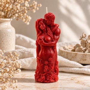 Puede incluir: Una vela de pilar de color rojo intenso con forma de dos figuras abrazadas, con la parte inferior decorada con detalles de rosas esculpidas. Una mecha blanca sobresale de la parte superior de la cabeza. La escultura de cera muestra a dos personas.