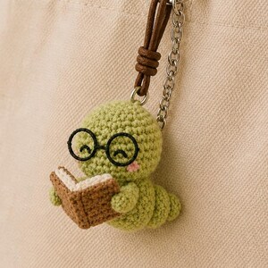 Puede incluir: Un gusano verde tejido a ganchillo con gafas leyendo un libro marrón, sujeto a un llavero. El llavero cuelga de un bolso tote beige. El gusano tiene mejillas rosadas y está hecho de hilo.