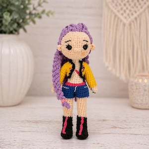 May include: A handmade crochet doll with purple hair in a braid, wearing a yellow jacket, blue shorts, and black boots with pink accents. The doll has a light skin tone and is standing on a wooden surface. The doll is approximately 15 cm tall.