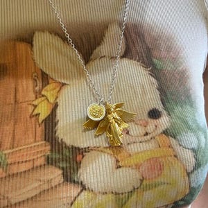 K&ouml;nnte beinhalten: Halskette mit silberner Kette und zwei Anh&auml;ngern: ein goldfarbener Engel mit ausgebreiteten Fl&uuml;geln und eine goldfarbene M&uuml;nze. Die Kette wird &uuml;ber einem hellen T-Shirt mit Hasenmotiv getragen.