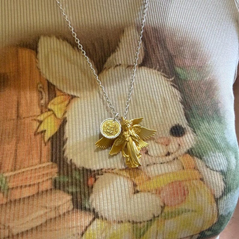K&ouml;nnte beinhalten: Halskette mit silberner Kette und zwei Anh&auml;ngern: ein goldfarbener Engel mit ausgebreiteten Fl&uuml;geln und eine goldfarbene M&uuml;nze. Die Kette wird &uuml;ber einem hellen T-Shirt mit Hasenmotiv getragen.