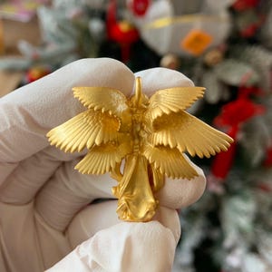 K&ouml;nnte beinhalten: Eine goldfarbene Engelsfigur mit detaillierten, geschichteten Fl&uuml;geln. Die Figur wird in einer behandschuhten Hand gehalten, mit einem verschwommenen Hintergrund eines dekorierten Weihnachtsbaums.