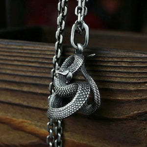May include: A silver snake pendant on a necklace. The pendant is a detailed sculpture of a coiled snake with textured scales. The necklace chain is silver and hangs from a wooden surface.