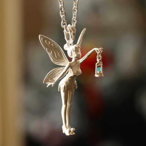 May include: A silver-colored pendant necklace featuring a fairy holding a lantern with a blue gem. The fairy has detailed wings and is suspended from a delicate chain. The pendant is a whimsical piece of jewelry.