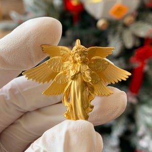 K&ouml;nnte beinhalten: Eine goldfarbene Engelsfigur mit ausgebreiteten Fl&uuml;geln und gefalteten H&auml;nden. Der Engel tr&auml;gt ein flie&szlig;endes Gewand und hat detaillierte Gesichtsz&uuml;ge. Die Figur wird in behandschuhten H&auml;nden gehalten, mit einem verschwommenen Hintergrund eines geschm&uuml;ckten Weihnachtsbaums.