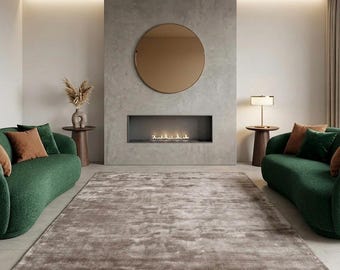 May include: A modern living room with a large, neutral-toned rug, two green curved sofas with brown and green pillows, and a fireplace. A round mirror hangs above the fireplace. Two small wooden side tables with lamps and vases are also present.