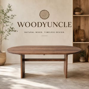 Puede incluir: Una mesa de comedor de madera marrón oscuro con tablero ovalado y dos patas verticales robustas conectadas por una viga de soporte horizontal. La pared detrás muestra el texto WOODYUNCLE con un icono de anillo de árbol y NATURAL WOOD. TIMELESS DESIGN.