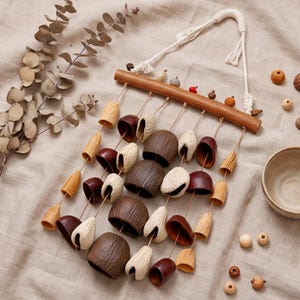 Peut inclure: Un carillon éolien mural composé d'une tige en bois horizontale suspendue par une corde blanche. Le carillon comprend plusieurs rangées d'éléments suspendus, dont des cloches en bois brun foncé, des pièces texturées beige clair et des perles.