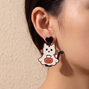 Könnte beinhalten: Ein Hängeohrring mit einem schwarzen herzförmigen Stecker, der mit einer weißen Katzenfigur verbunden ist, die ein Geisterkostüm trägt und einen orangefarbenen Kürbiseimer hält. Die Katze hat schwarze Schnurrhaare, einen schwarzen Schwanz und ein lächelndes Gesicht.