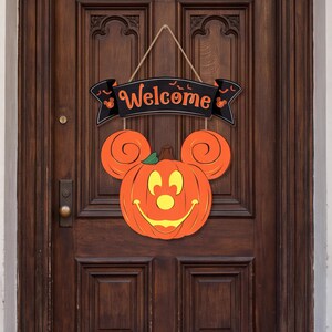 Könnte beinhalten: Ein hängendes Türschild mit einem schwarzen Banner, dem orangefarbenen Schriftzug Welcome und kleinen Fledermaussilhouetten. Darunter befindet sich ein großer orangefarbener Kürbis in Form eines Mauskopfes mit lächelndem Gesicht und gelben Augen.