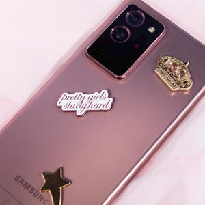 May include: A rose-gold Samsung phone with a gold crown charm, a gold star charm, and a white sticker that reads "pretty girls study hard." The phone's camera is visible at the top.