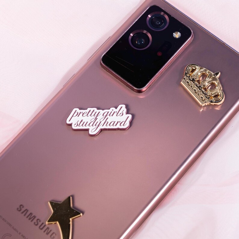May include: A rose-gold Samsung phone with a gold crown charm, a gold star charm, and a white sticker that reads "pretty girls study hard." The phone's camera is visible at the top.