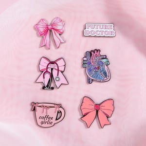 May include: A collection of pink enamel pins. Designs include a bow, a bow with a stethoscope, a heart, a coffee cup with "coffee girlie" text, and a pin with "FUTURE DOCTOR" text.