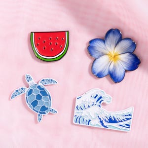 May include: Four enamel pins with tropical themes. A watermelon slice with red and green colors, a blue and white flower, a blue sea turtle, and a white and blue wave design.