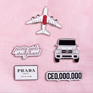 May include: A collection of five enamel pins featuring a white airplane with red accents, a white SUV, a white rectangular sign with the text "pretty girls study hard", a white rectangular sign reading "PRADA MARFA 1837 MI", and a sign reading "CEO,000,000".