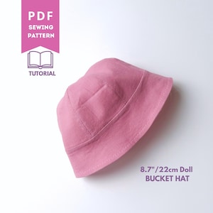Op de afbeelding: Een roze emmerhoed voor een pop, voorzien van een gestikte rand en kroon. De afbeelding bevat de tekst PDF SEWING PATTERN TUTORIAL en 8.7"/22cm Doll BUCKET HAT, die de maat van het voltooide item in inches en centimeters aangeeft.