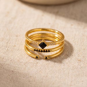 May include: A gold-toned multi-band ring featuring a central square-cut black stone flanked by small clear stones, a second band with a row of tiny black stones, and a third open-ended band accented with two small black stones at the terminals.