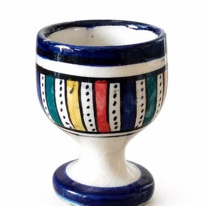 Puede incluir: Un huevero de cerámica pintado a mano con base blanca y borde azul. El cuerpo del cuenco presenta rayas verticales en varios colores, incluyendo azul, verde, amarillo y rojo, separadas por líneas de puntos. El huevero tiene una base de pedestal.