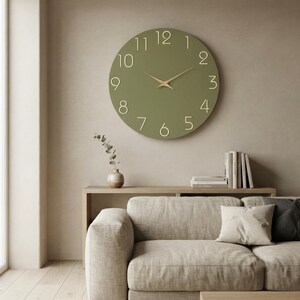 May include: A large, circular wall clock with a matte olive green face and raised, light-colored numerals from one to twelve. The clock features two slender, pointed hands in a light wood tone positioned at the center of the circular dial.