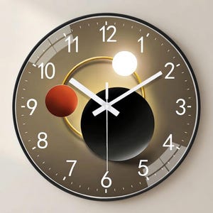 May include: A round wall clock with a black frame and a brown gradient face. The dial features white Arabic numerals from 1 to 12, white hour, minute, and second hands, and a graphic design of a large black circle, a red circle, and a white circle.