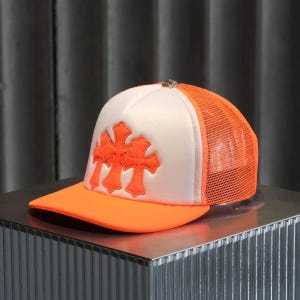 Puede incluir: Gorra de camionero con panel frontal blanco y visera y parte trasera de malla naranja. El sombrero presenta dos diseños de cruces naranjas en la parte delantera y un pequeño adorno plateado en la parte superior.