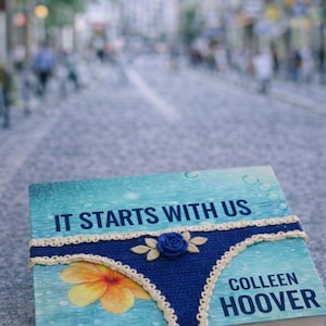 Könnte beinhalten: Ein Buch mit dem Titel "IT STARTS WITH US" von Colleen Hoover. Das Buch ist mit einem blauen Häkeldesign verziert, das Unterwäsche ähnelt, verziert mit einer blauen Rose und einer gelben Blume. Der Hintergrund ist eine verschwommene Stadtstraße.