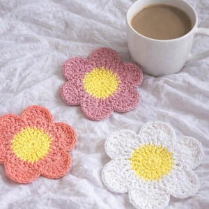 May include: Three handmade crochet flower coasters in shades of pink, peach, and white, each with a yellow center. A white mug filled with coffee sits nearby. The coasters are on a white, textured surface.