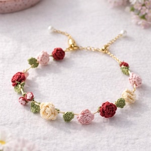May include: A delicate, handmade bracelet featuring crocheted flowers in shades of red, pink, and cream, interspersed with green leaves. The bracelet is strung on a gold-colored chain, with a total length of approximately 7 inches.