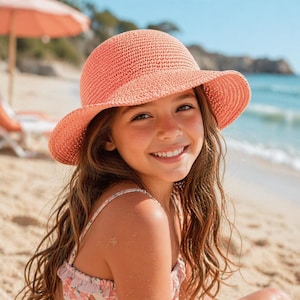 May include: A young person wearing a coral-coloured sun hat with a wide brim, smiling on a sandy beach. The hat is made of woven material. The background includes the ocean, a beach umbrella, and a sunny sky.