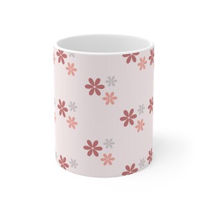 Könnte beinhalten: Weißer Keramikbecher mit hellrosa Hintergrund und einem sich wiederholenden Blumenmuster. Die Blumen sind in verschiedenen Rot-, Rosa- und Grautönen gehalten. Das Blumendesign ist einfach, mit abgerundeten Blütenblättern.