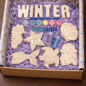 May include: A craft kit in a cardboard box filled with light purple paper shreds. It contains white plaster figures of a seahorse, starfish, seashell, mermaid tail, turtle, and jellyfish, plus paint pots, a brush, glitter, and blue gems.