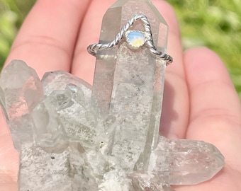 May include: A silver ring with a twisted band and a small, iridescent opal gemstone. The ring is displayed on a cluster of clear quartz crystals.