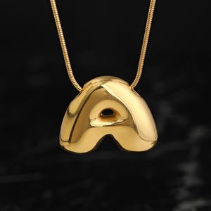 May include: A gold-toned necklace featuring a polished, puffed letter A pendant suspended from a matching snake chain. The pendant has a smooth, rounded, and reflective surface with a distinct three-dimensional shape and a central cutout.