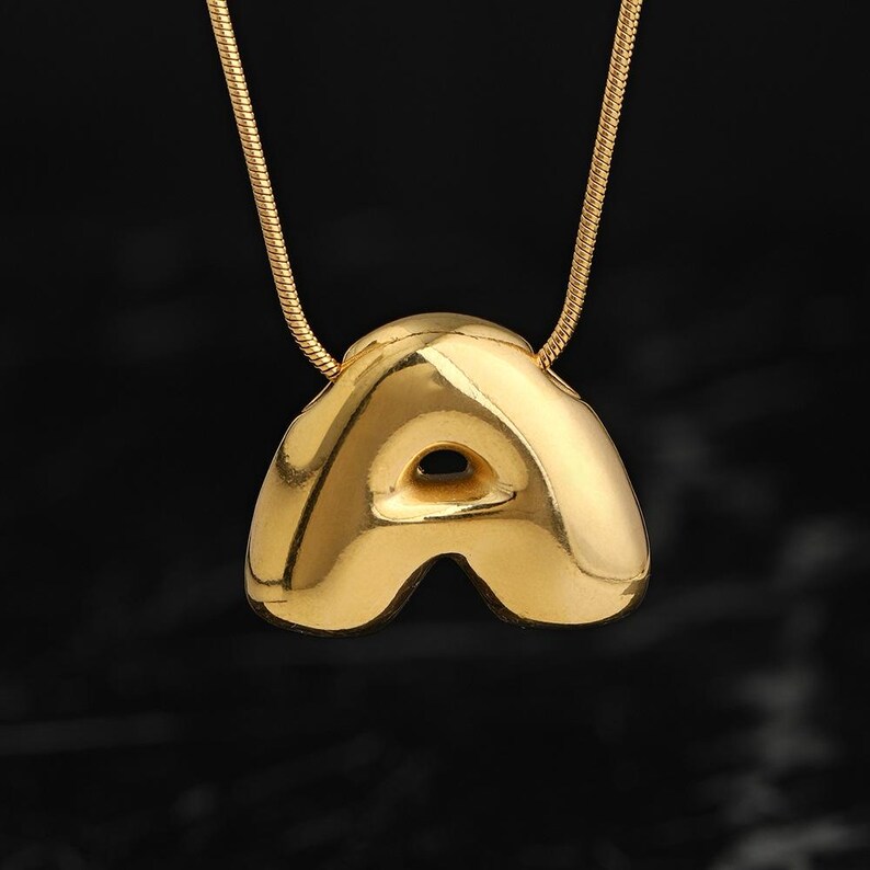 May include: A gold-toned necklace featuring a polished, puffed letter A pendant suspended from a matching snake chain. The pendant has a smooth, rounded, and reflective surface with a distinct three-dimensional shape and a central cutout.